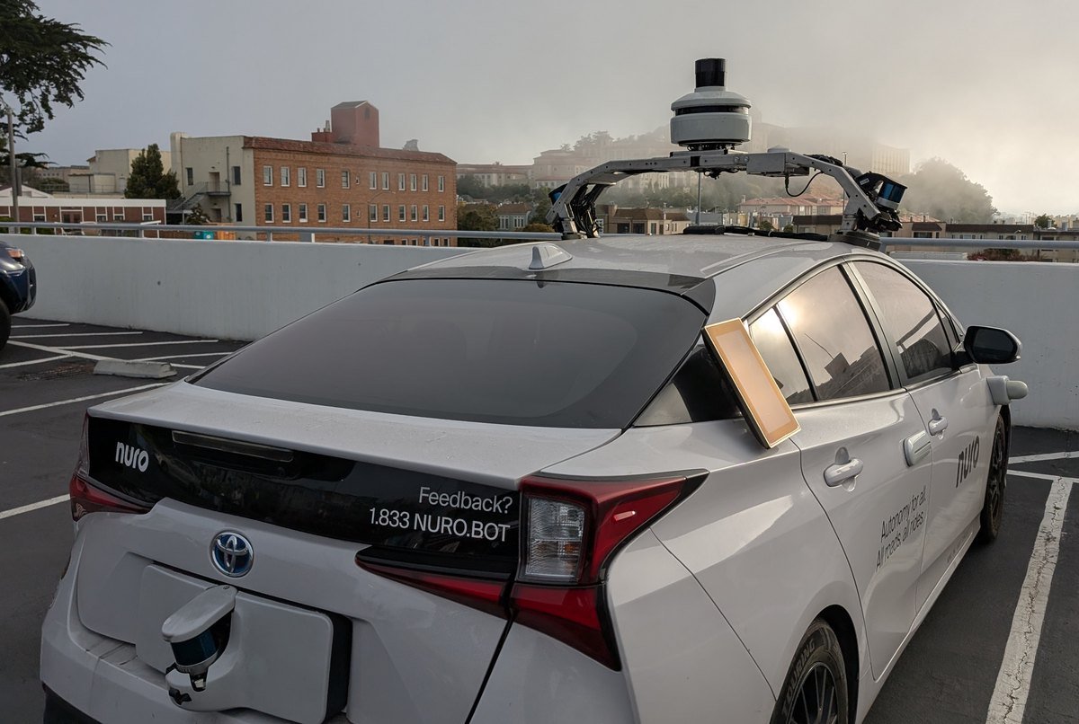Rear view of a white Nuro test car with a roof-mounted sensor rack, parked on a rooftop lot in San Francisco on a foggy day.