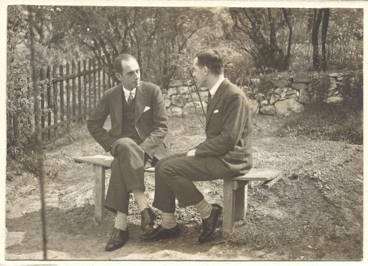 An old sepia family album photograph of two men in suits sitting on a wooden bench in a garden, facing each other in conversation