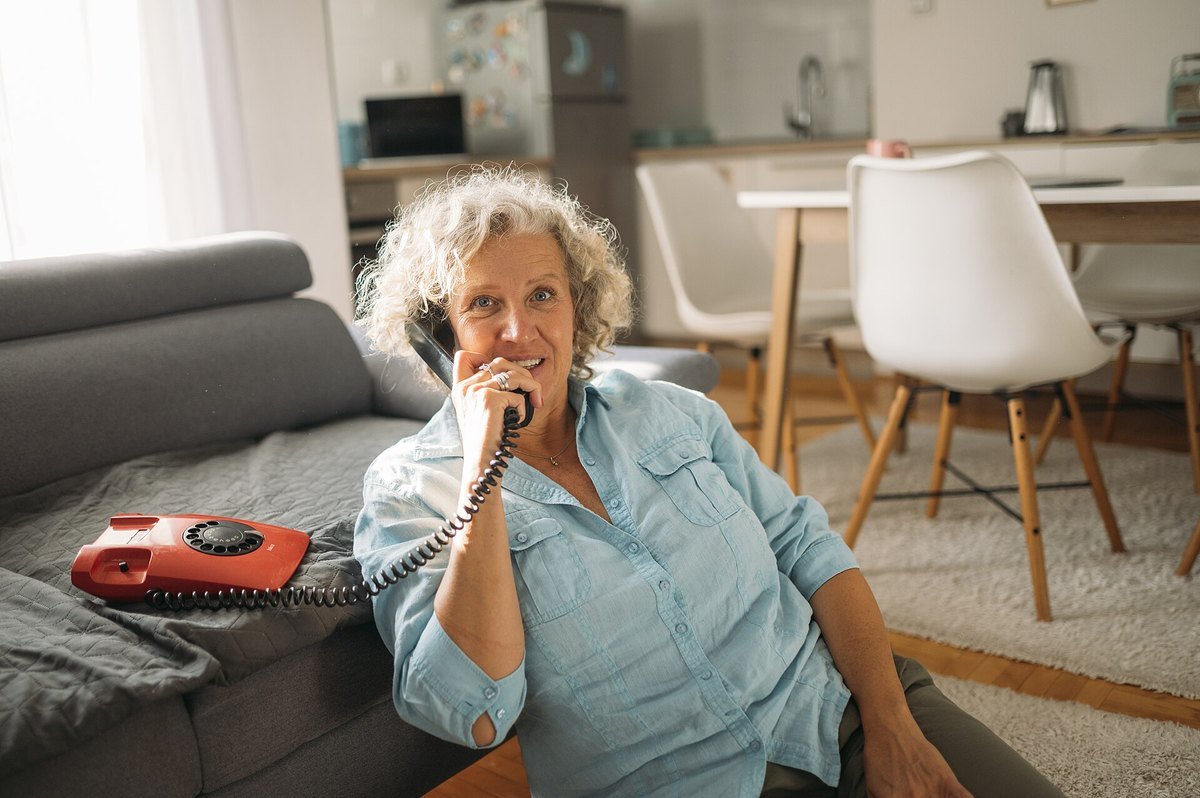 An older woman sits on the floor by a sofa, talking on a red rotary telephone