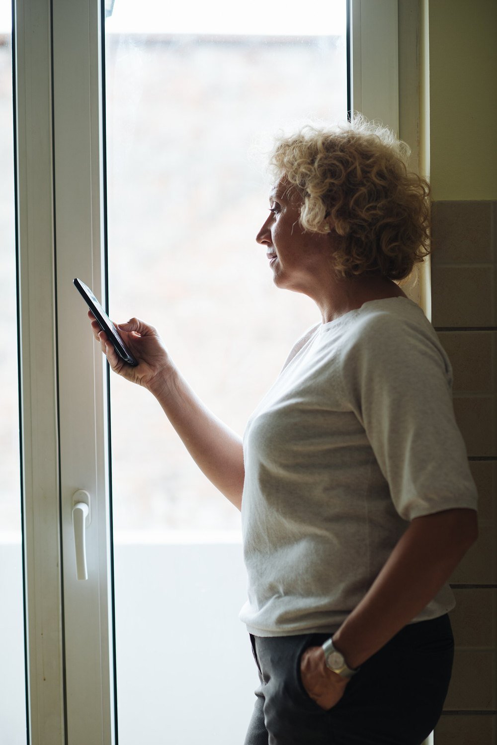 An older woman with curly hair stands by a window, reading something on her smartphone