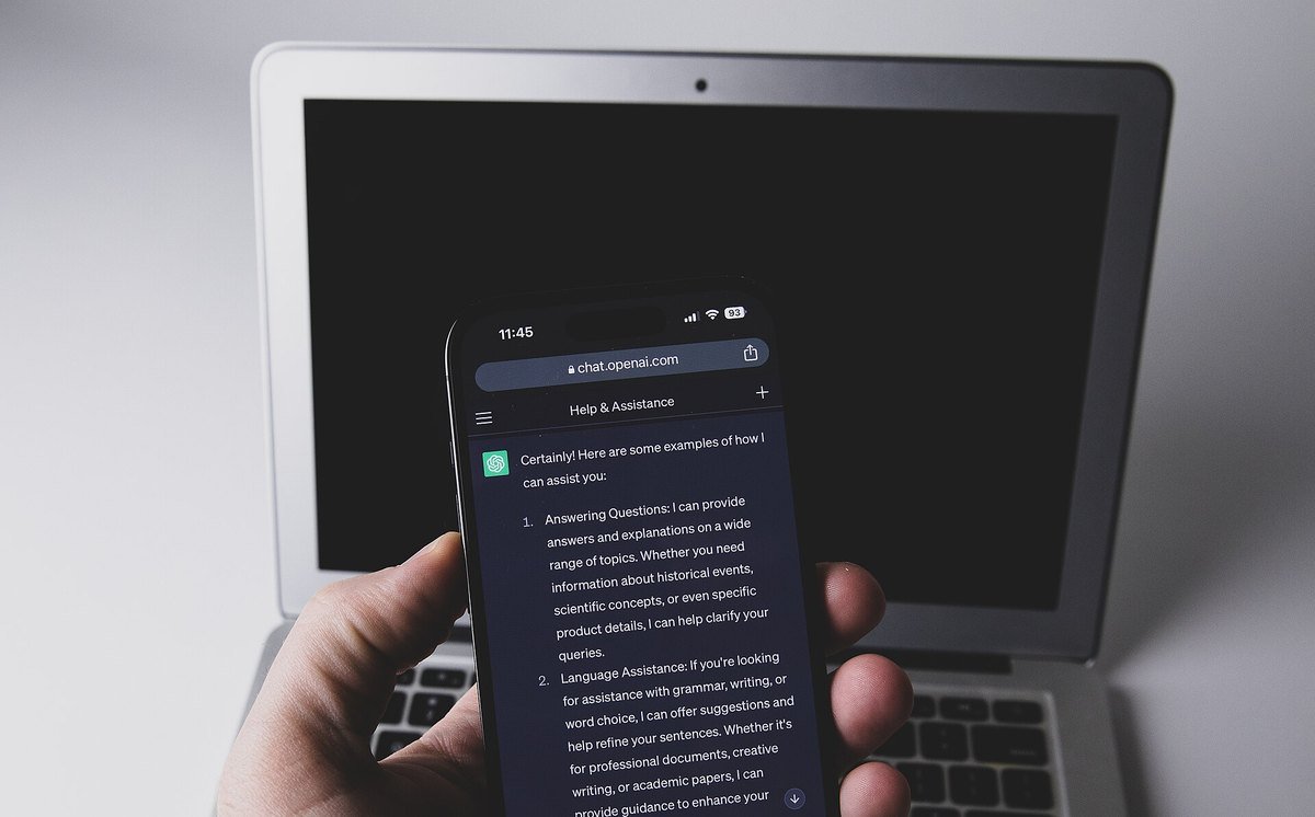 A hand holds a smartphone showing a ChatGPT conversation on chat.openai.com in dark mode, in front of an open laptop
