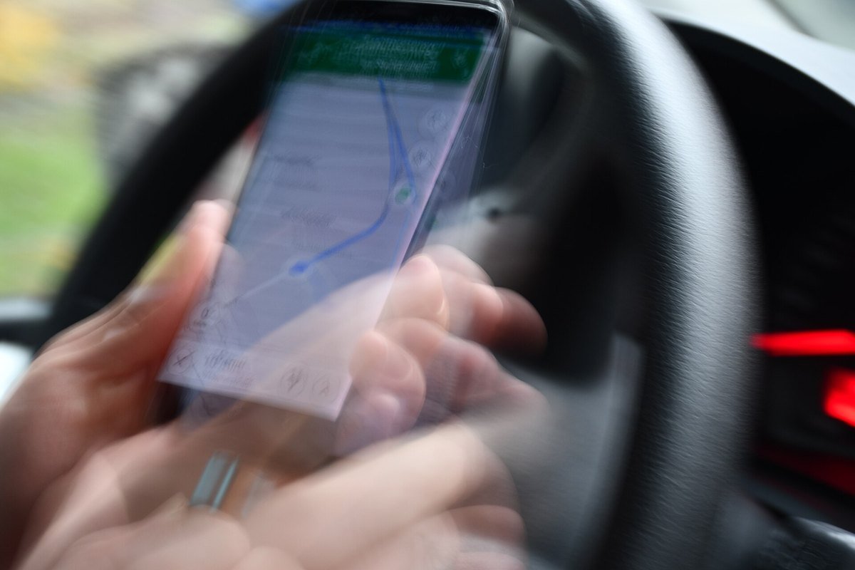 A blurred photo of a hand holding a smartphone showing a map in front of a car steering wheel