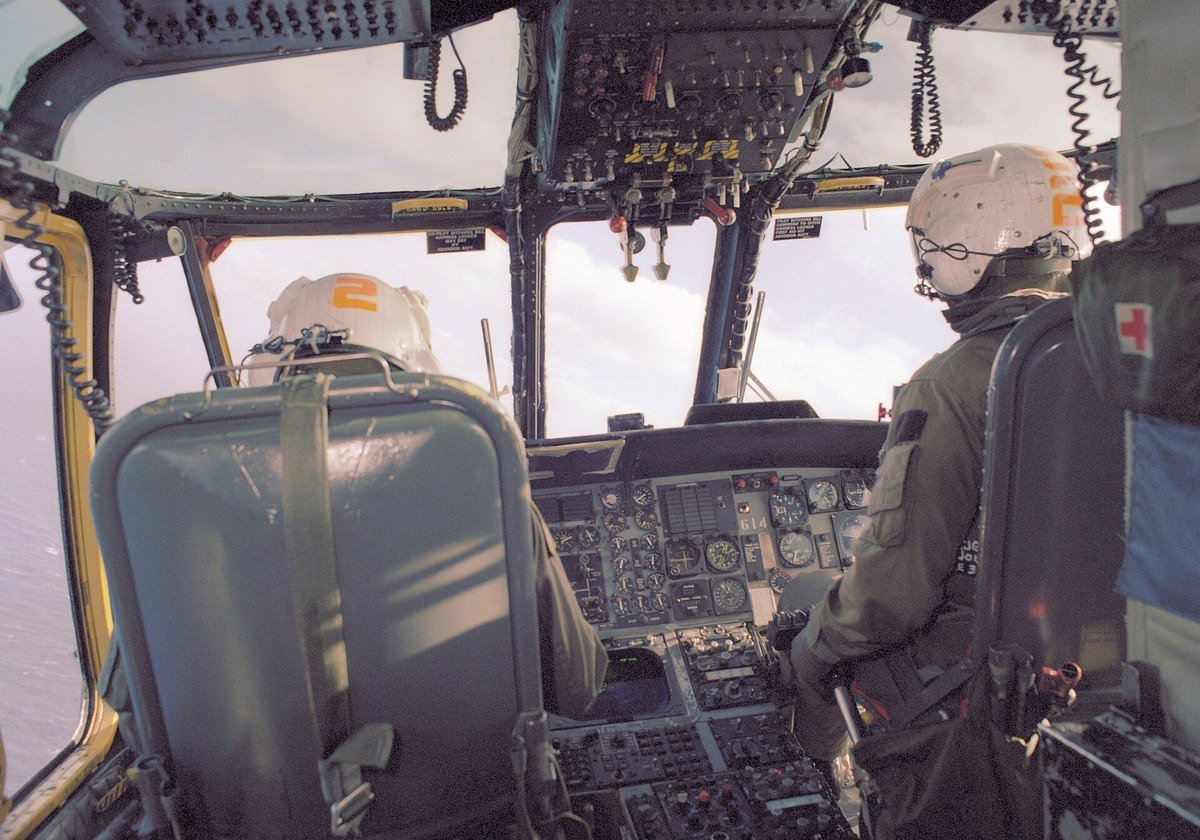 Two crew members in helmets sit side by side in a helicopter cockpit, one in the pilot seat and one in the copilot seat