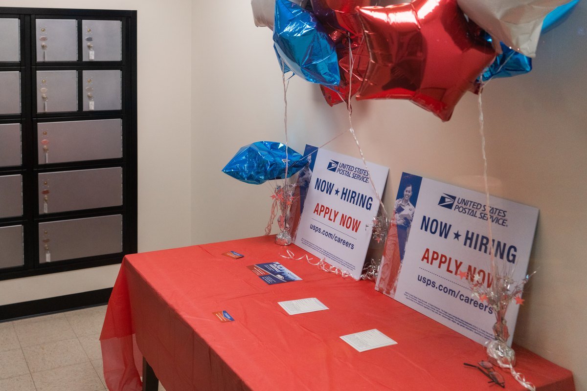 A red-covered table in a post office lobby with two United States Postal Service Now Hiring Apply Now signs, red, white and blue star balloons, and rows of mailboxes on the left wall.