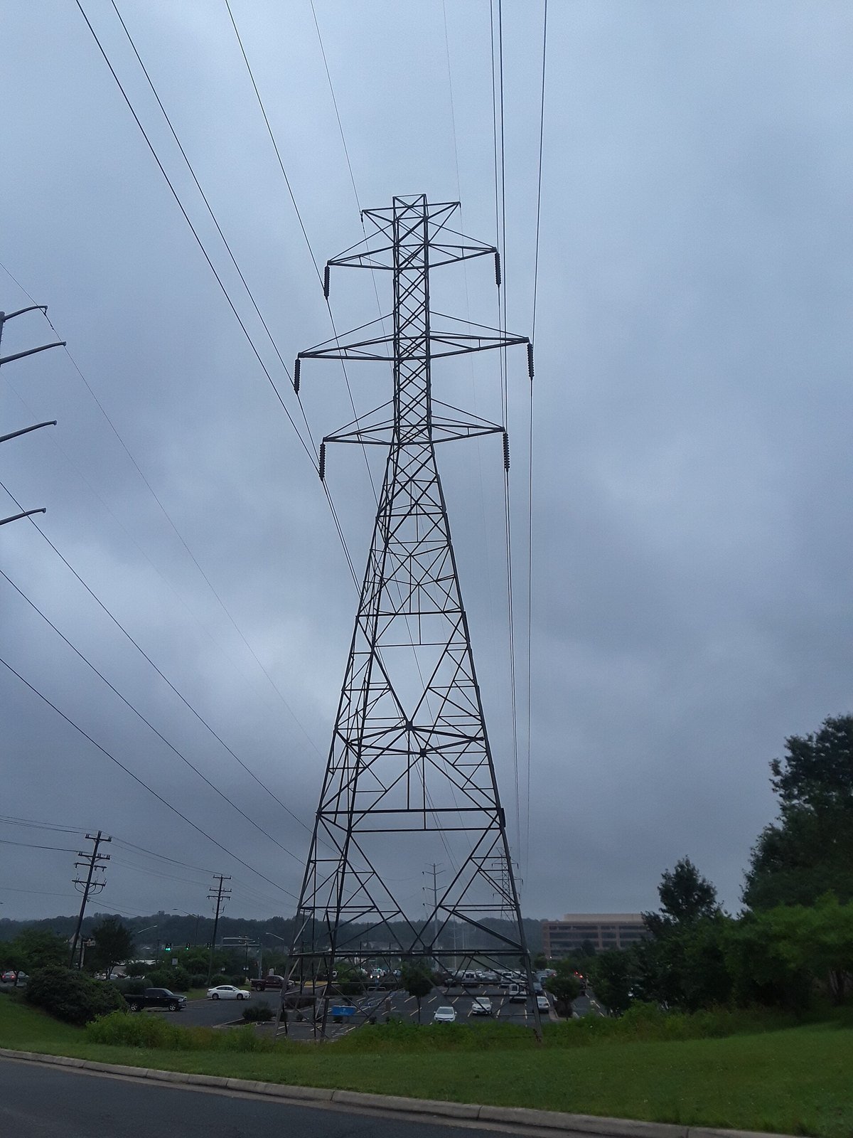 A tall steel electricity transmission tower carrying high-voltage lines over a cleared corridor under a gray sky in Kingstowne, Northern Virginia.