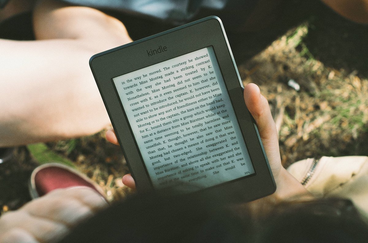 Two hands holding an older Kindle e-reader outdoors on the grass, showing a page of text