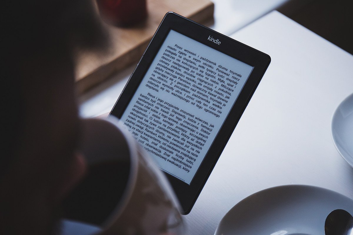 A hand holding an older Kindle e-reader at a cafe table next to a coffee cup