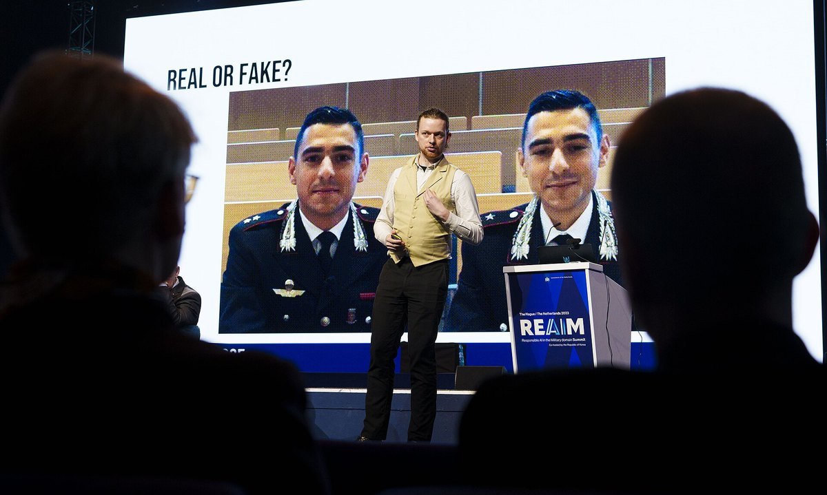 Audience silhouettes in front of a large screen with the words Real or Fake above a photo of three men