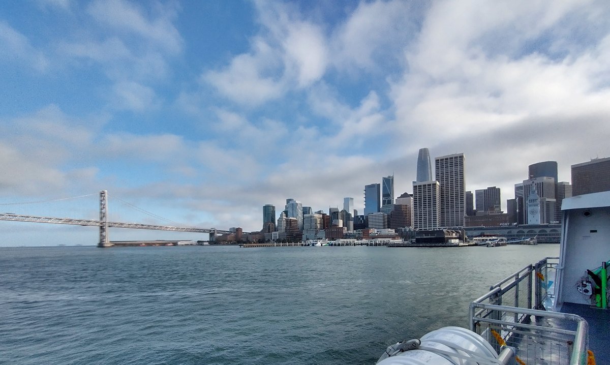 San Francisco skyline seen from a ferry on the bay, with the Bay Bridge on the left and the South of Market and downtown towers under a cloudy sky.