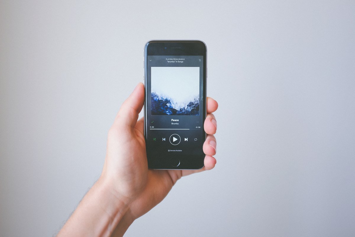 A hand holding a smartphone with the Spotify Now Playing screen open, showing a song with play and skip buttons