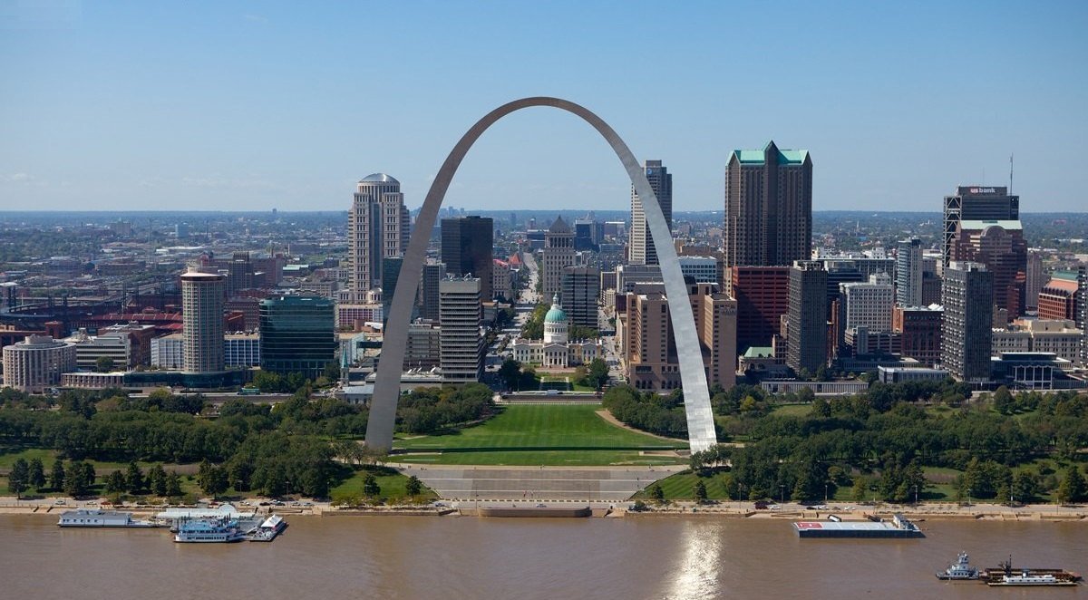 Aerial view of the Gateway Arch in St. Louis framing the downtown skyline, with riverboats on the brown Mississippi River in front.