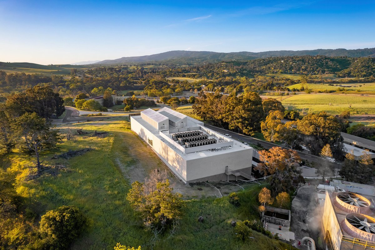 Aerial view at sunrise of the white, flat-roofed Stanford Research Computing Facility on the SLAC campus, surrounded by green hills and oak trees, with large cooling fans visible in the foreground.