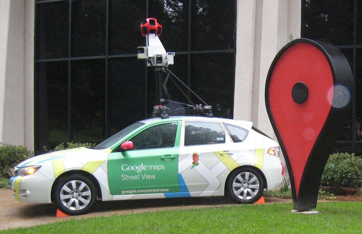 A Google Maps Street View car with a camera mast on its roof parked on a lawn next to a giant red map pin sculpture