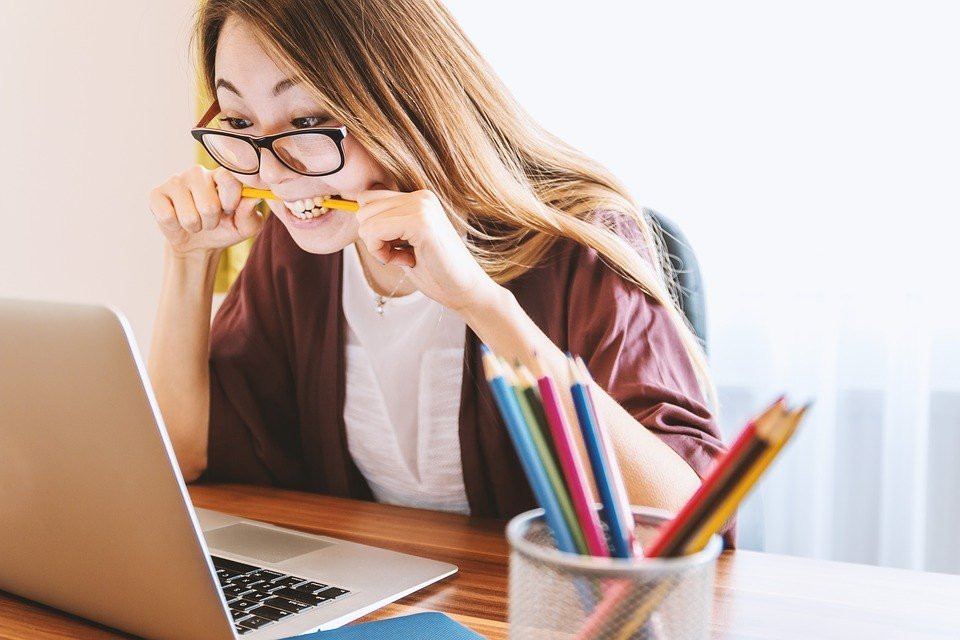 A young woman in glasses bites a pencil while looking at an open laptop on a desk next to a cup of colored pencils