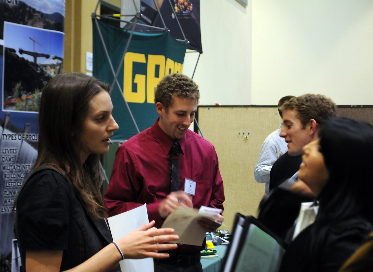 Engineering students in business clothes hold resumes and talk with a recruiter at a career fair table in a gym, with an employer banner behind them.