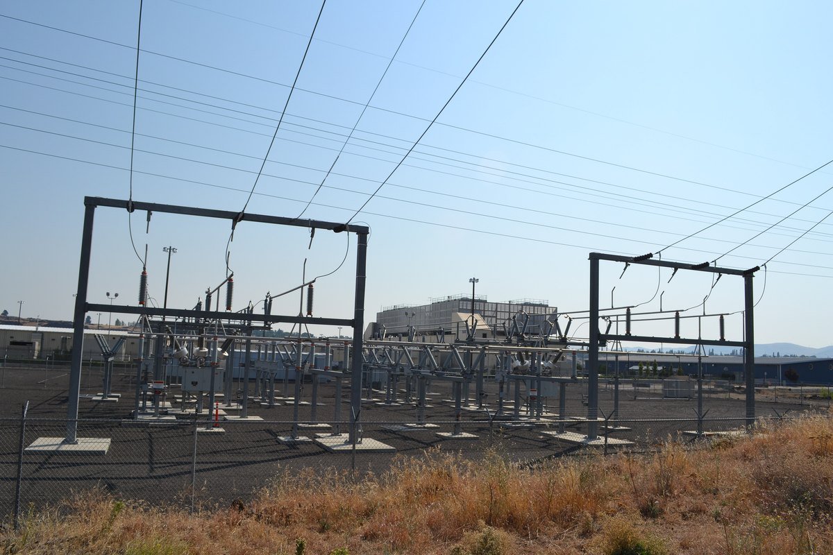 An electrical substation with steel frames, insulators and high-voltage wires in front of a large Google data center building in The Dalles, Oregon, under a clear sky.
