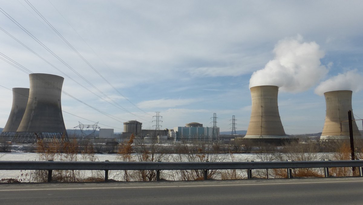 The cooling towers of the Three Mile Island nuclear plant in Pennsylvania, two of them releasing steam, seen across the Susquehanna River with power lines in the foreground.