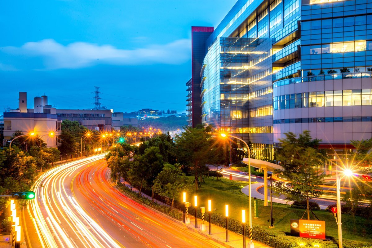 A modern glass-walled semiconductor fab building lit up at dusk beside a curving road with light trails from traffic in Taiwan.