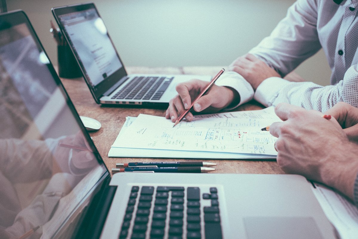 Two people working side by side at laptops on a shared desk, one writing notes on printed pages with a pencil.