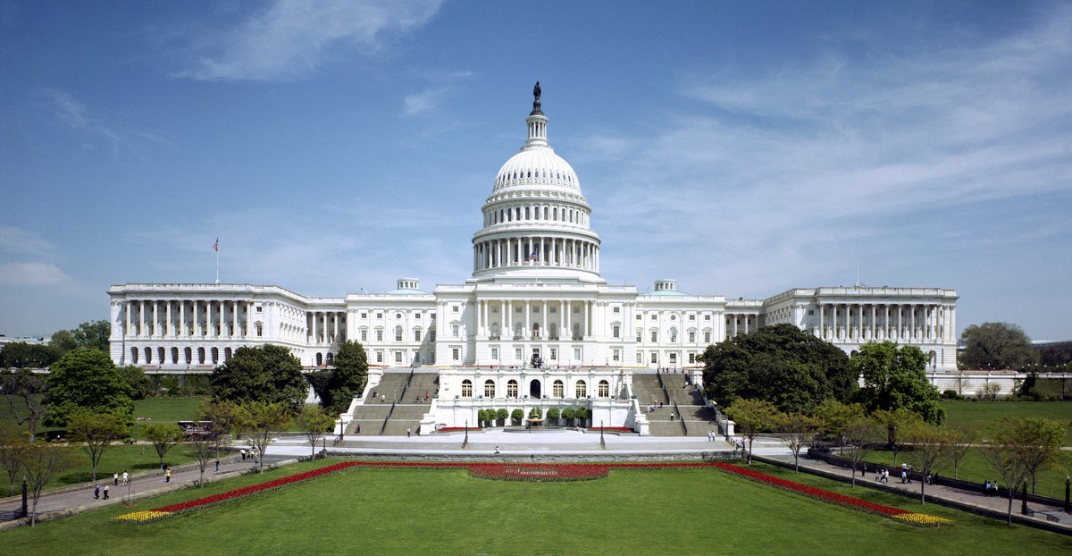 The west front of the United States Capitol in Washington, D.C., with its white dome, steps and lawn under a blue sky.