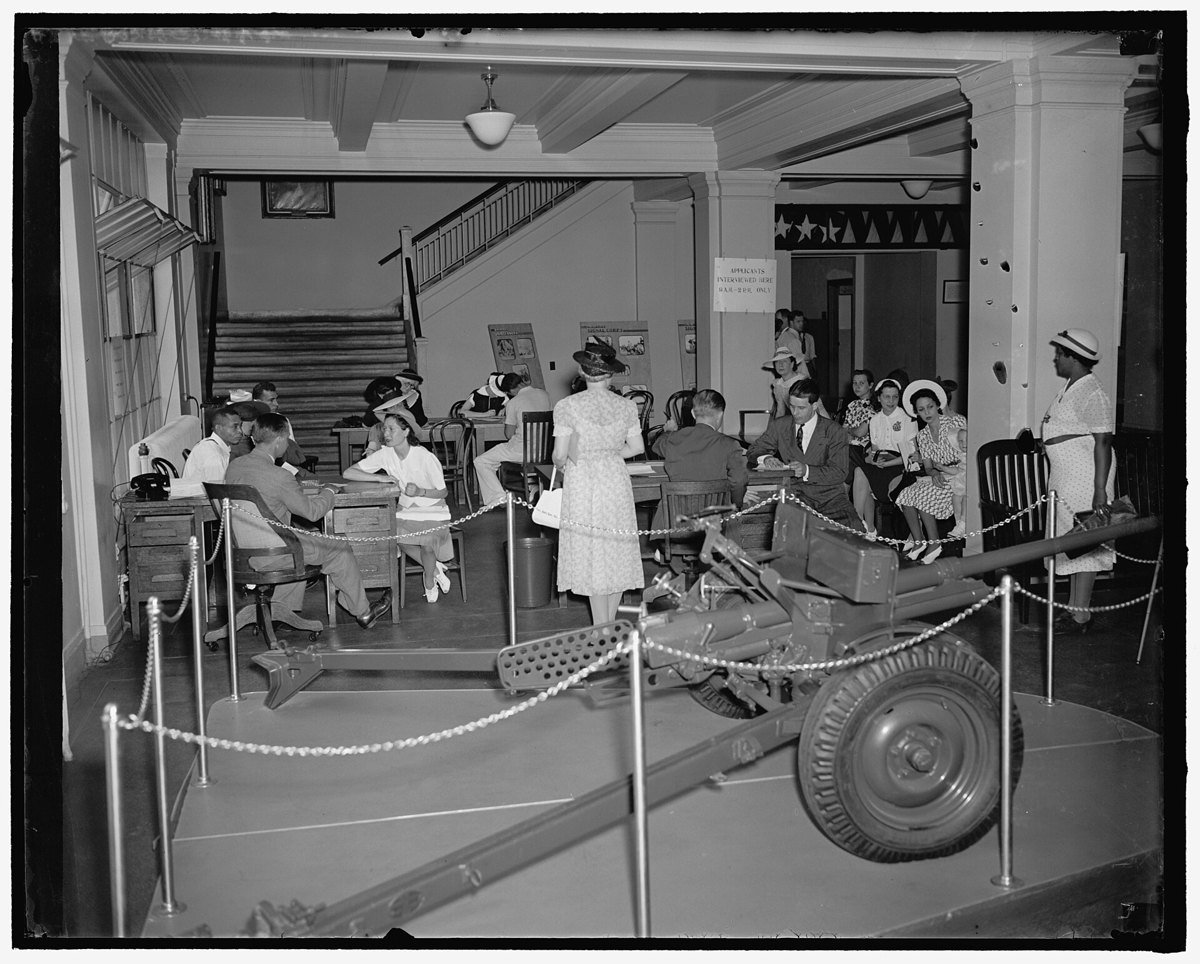 Black and white photograph of a crowded ground-floor hall in Washington in 1940 where job applicants sit at tables filling out forms and wait for interviews, with a field gun on display in the foreground.