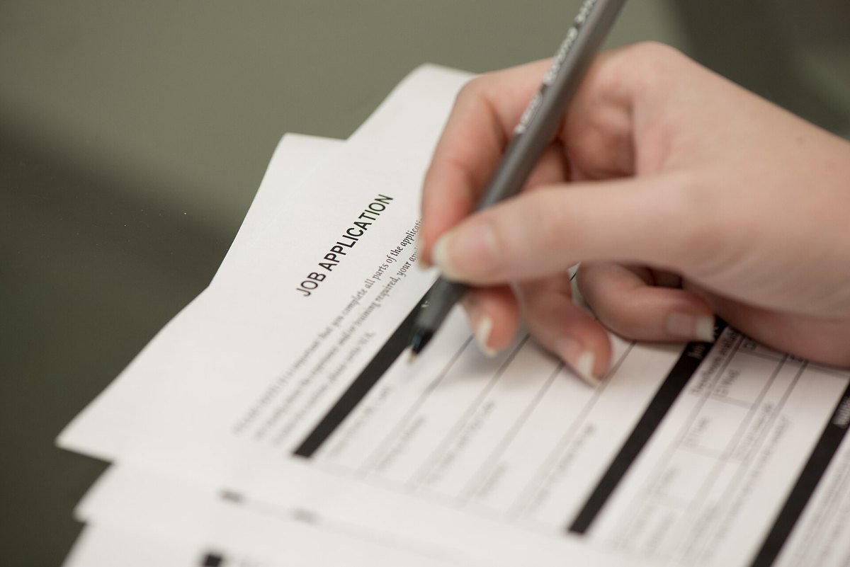 Close-up of a hand holding a black pen over a printed job application form, with more forms stacked underneath on a desk.