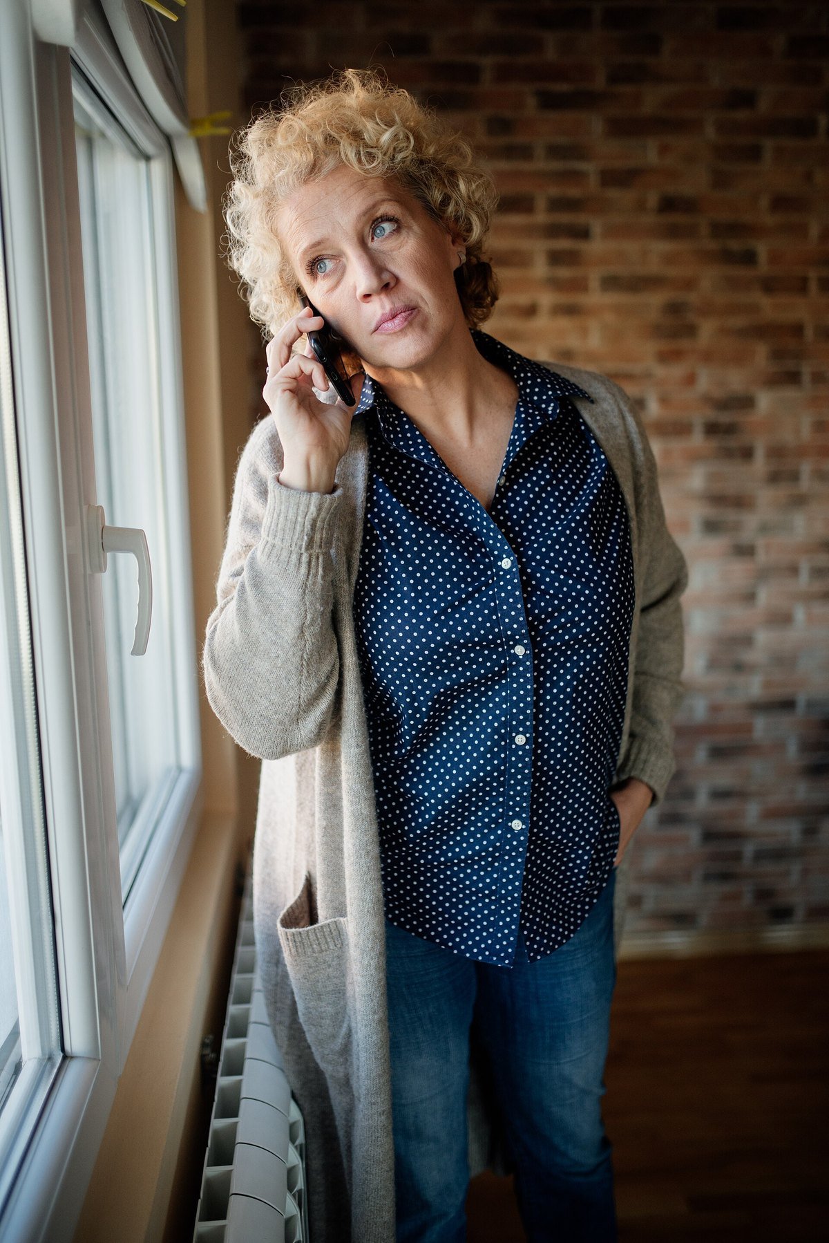 A woman with curly blond hair stands by a window, holding a cell phone to her ear with a worried look