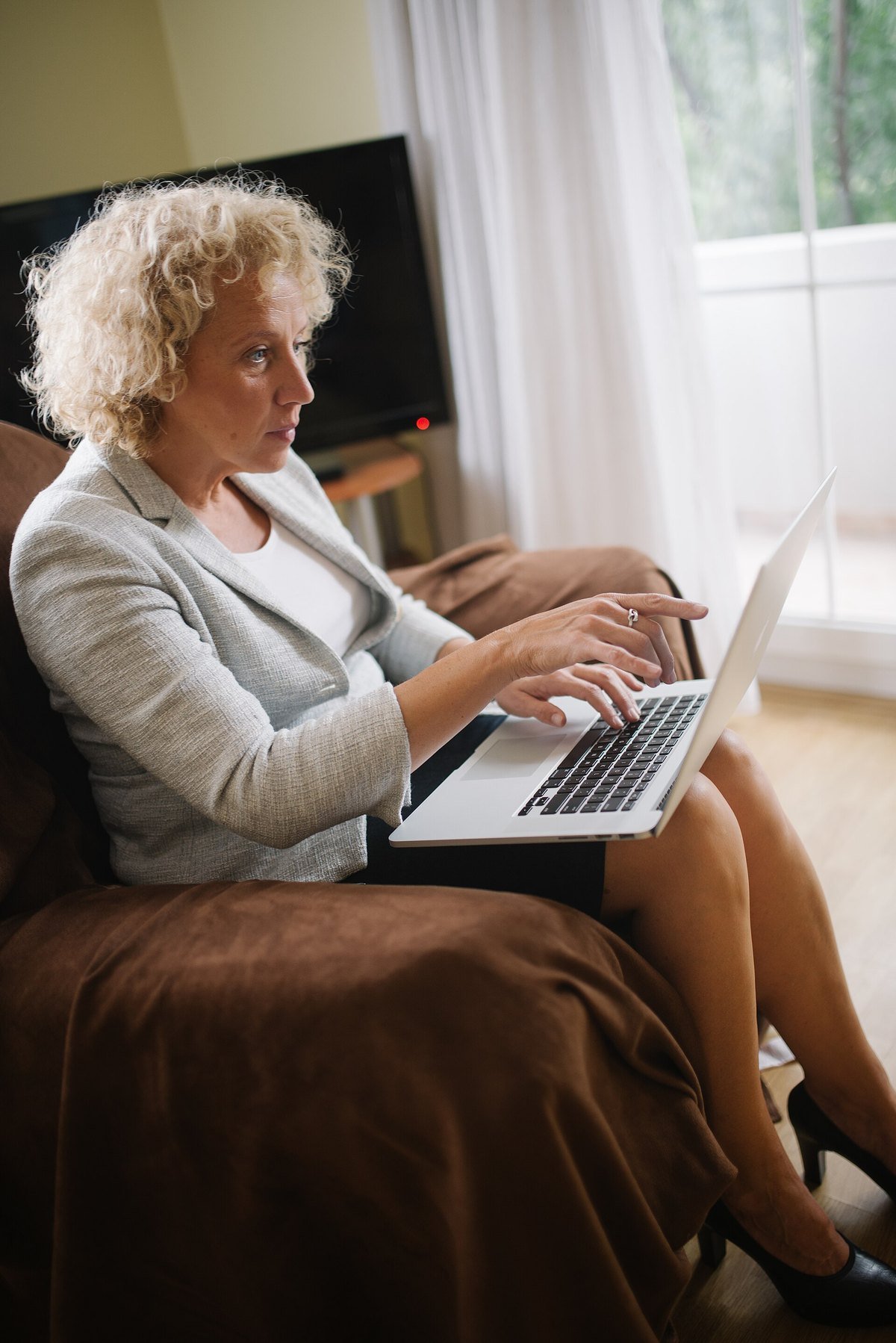 A woman with curly hair sits with a laptop on her lap and points at something on the screen with a puzzled look