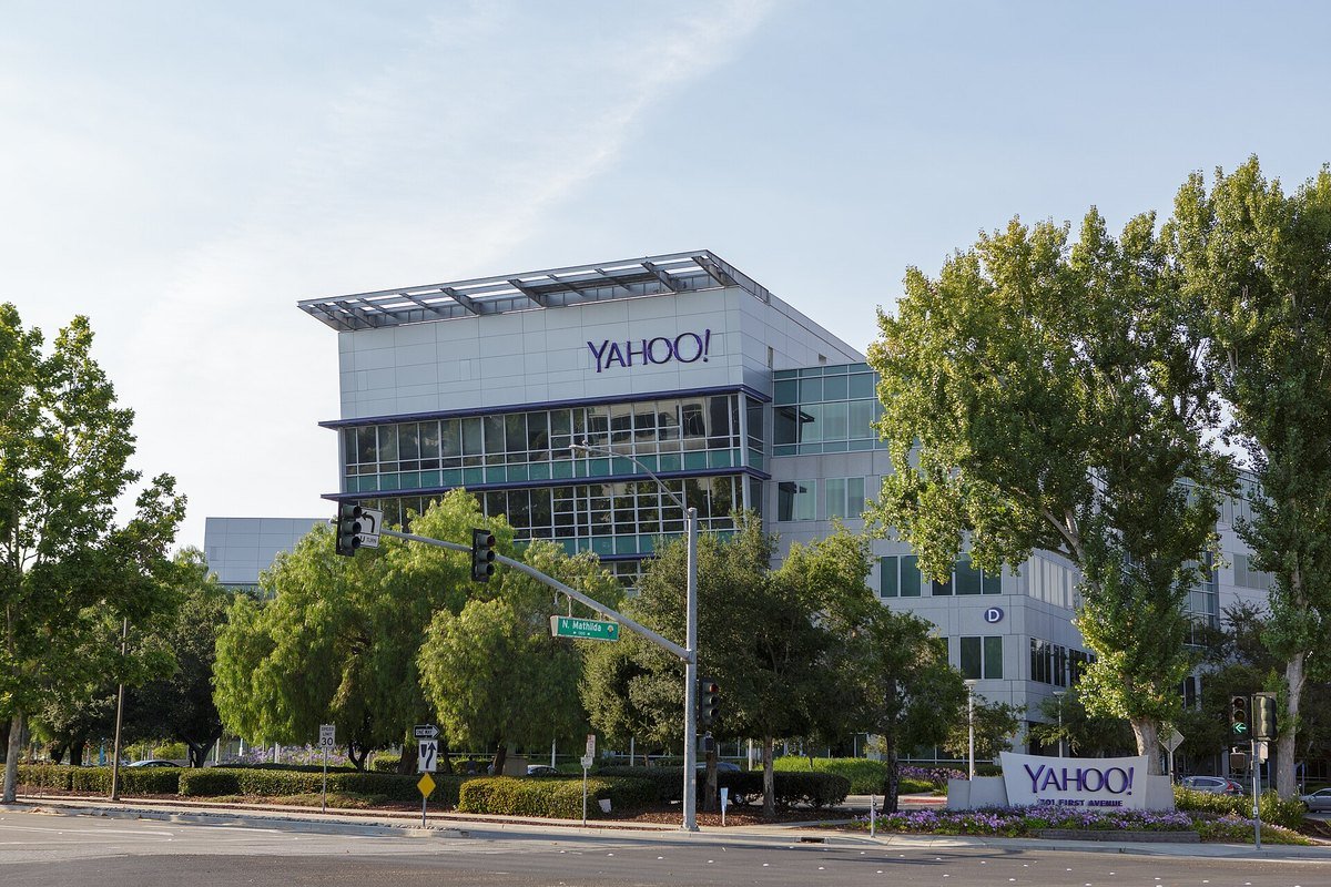 A glass and white office building with a purple Yahoo sign on the roof, behind trees and a street