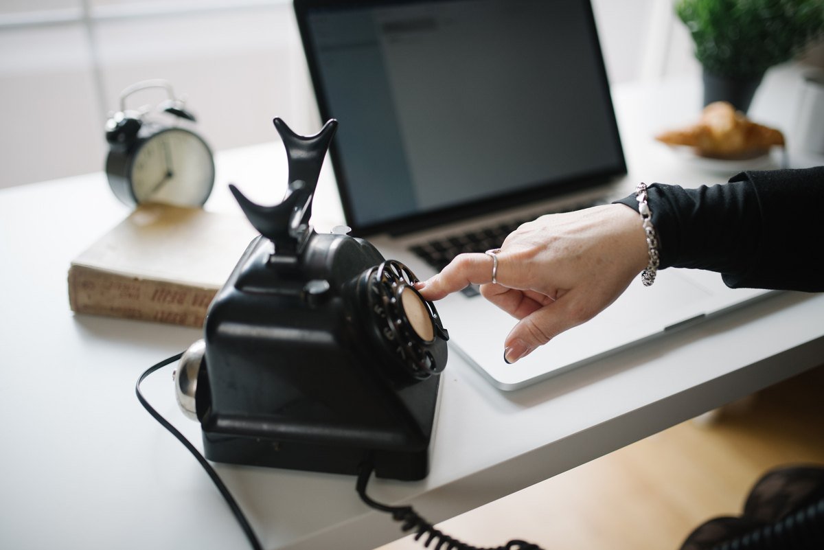 A hand dialing a black vintage rotary telephone on a white desk beside an open laptop and an alarm clock