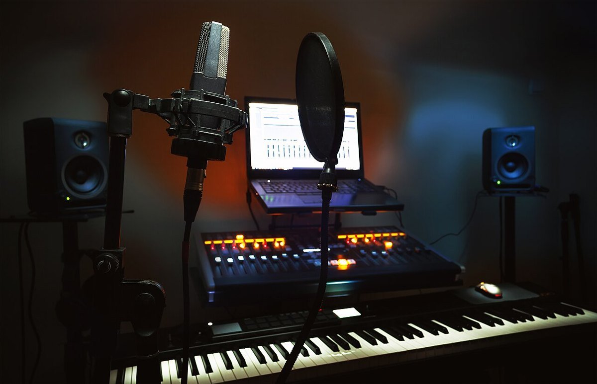 A small home recording setup in a dark room: a microphone with a pop filter on a stand, a laptop, a mixing desk with lit buttons, a keyboard and two speakers.