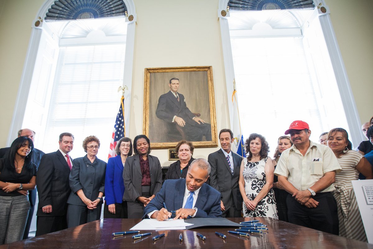 Massachusetts Governor Deval Patrick seated at a desk signing a bill, with a group of people standing behind him in a room with tall windows and a portrait