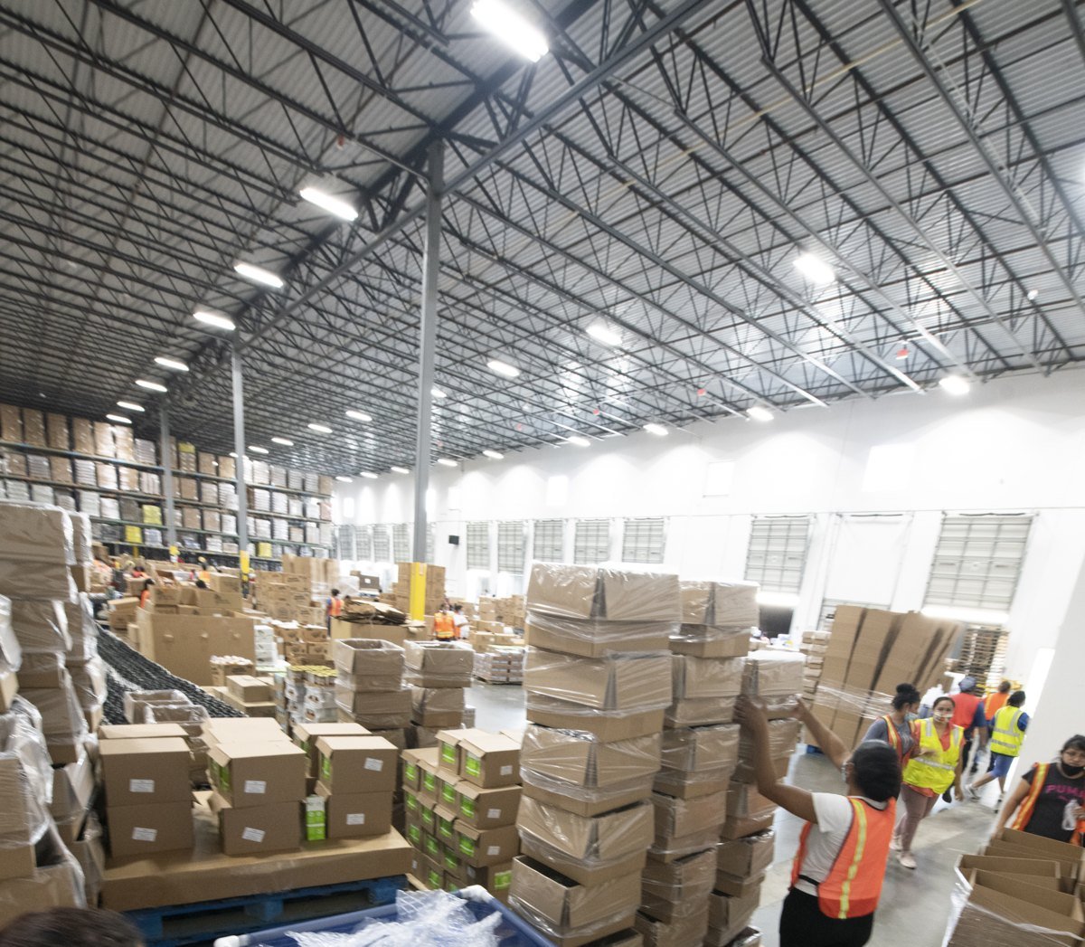 Wide view of a large warehouse with stacked cardboard boxes and workers in safety vests packing and moving boxes