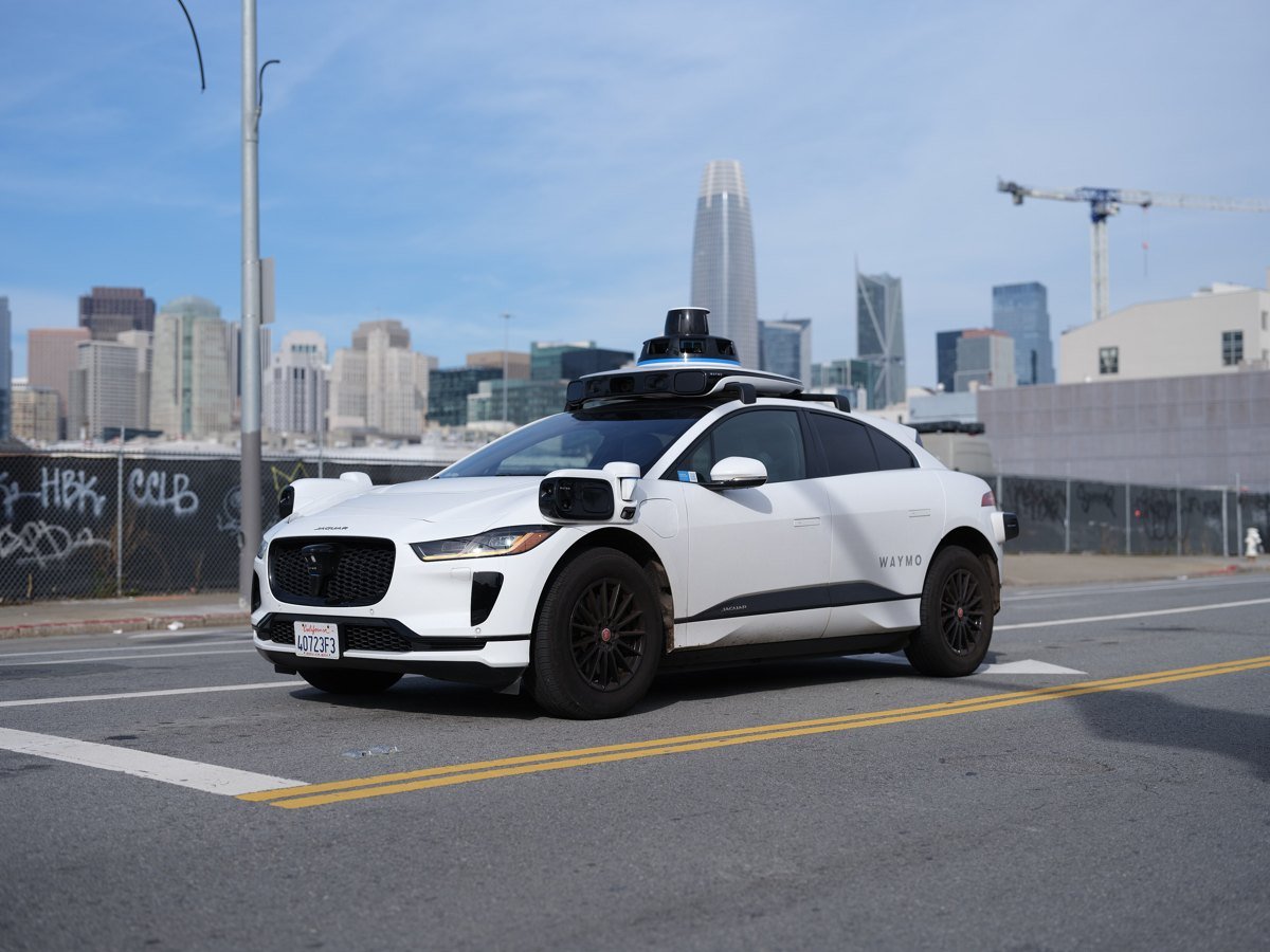 A white Waymo Jaguar I-Pace with roof-mounted sensors parked on a San Francisco street, with downtown towers and a construction crane behind it.